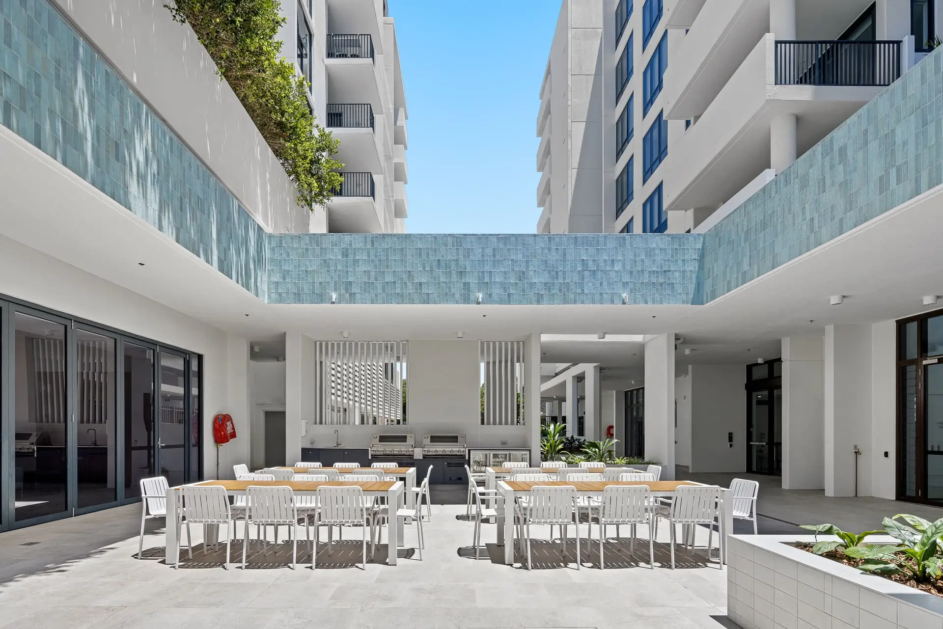 Outdoor dining area with white tables and chairs, nestled between modern apartment buildings in a Gold Coast retirement community. A barbecue grill and countertop are in the background, with lush plants and blue sky overhead.