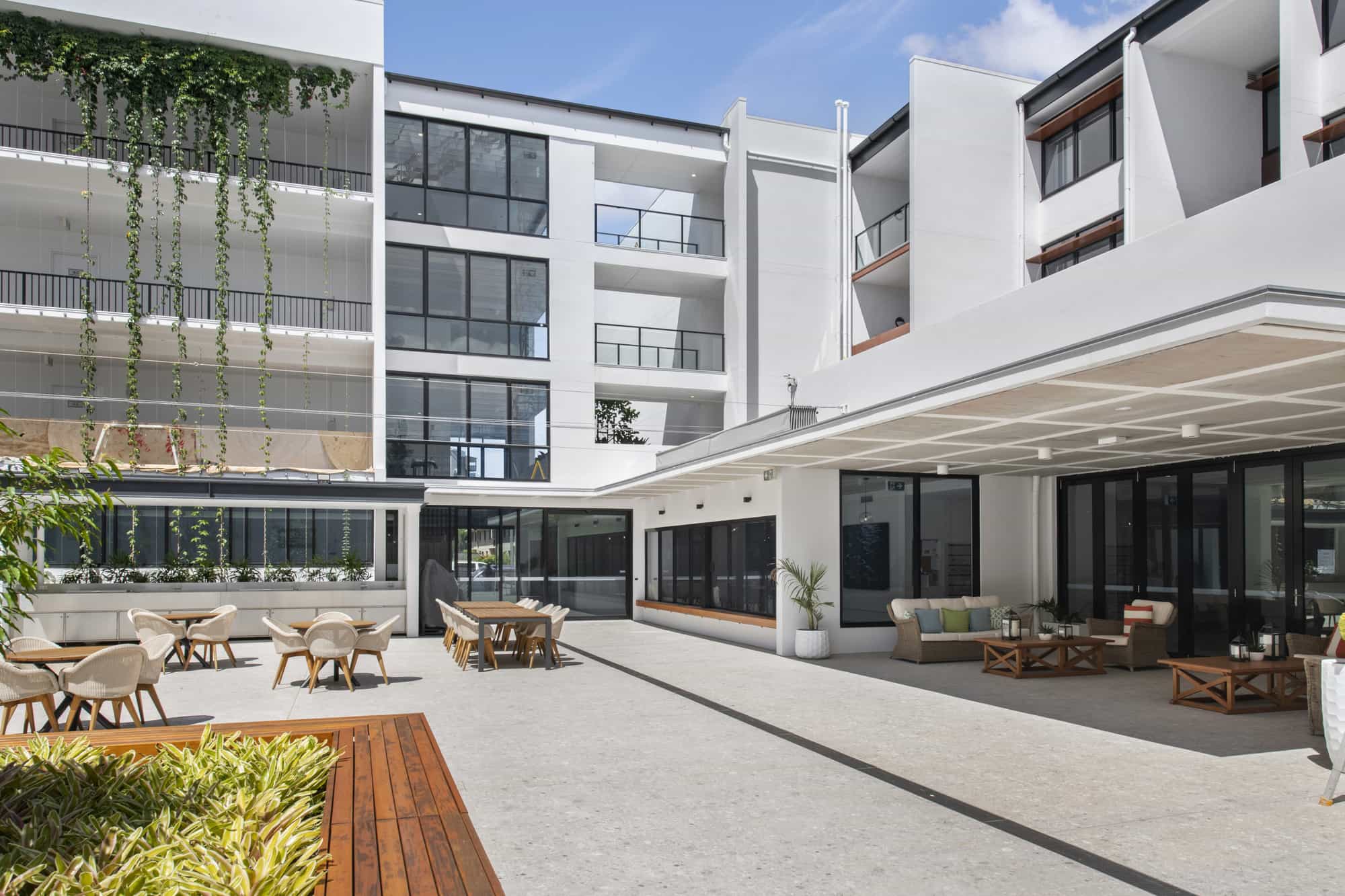 Modern open-air courtyard of a Sunshine Coast Retirement Community, featuring outdoor seating, tables, lush greenery, and large glass windows. The area is bright and sunny, with white walls and a contemporary architectural design.