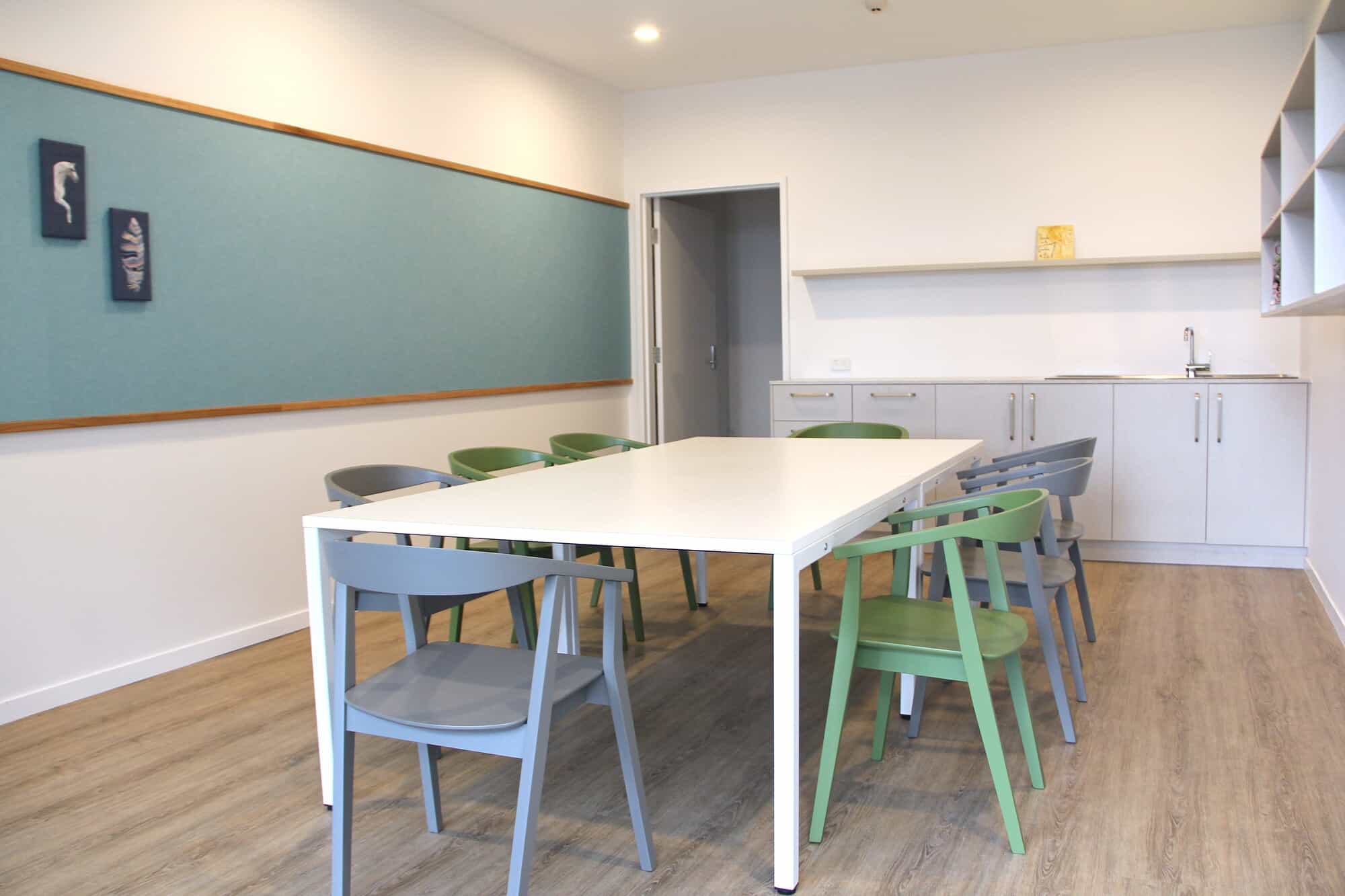 A modern, minimalist room in a Sunshine Coast Retirement Community features a white table with gray and green chairs, a blue bulletin board, wooden flooring, and a small kitchenette with cabinets and a sink in the background.