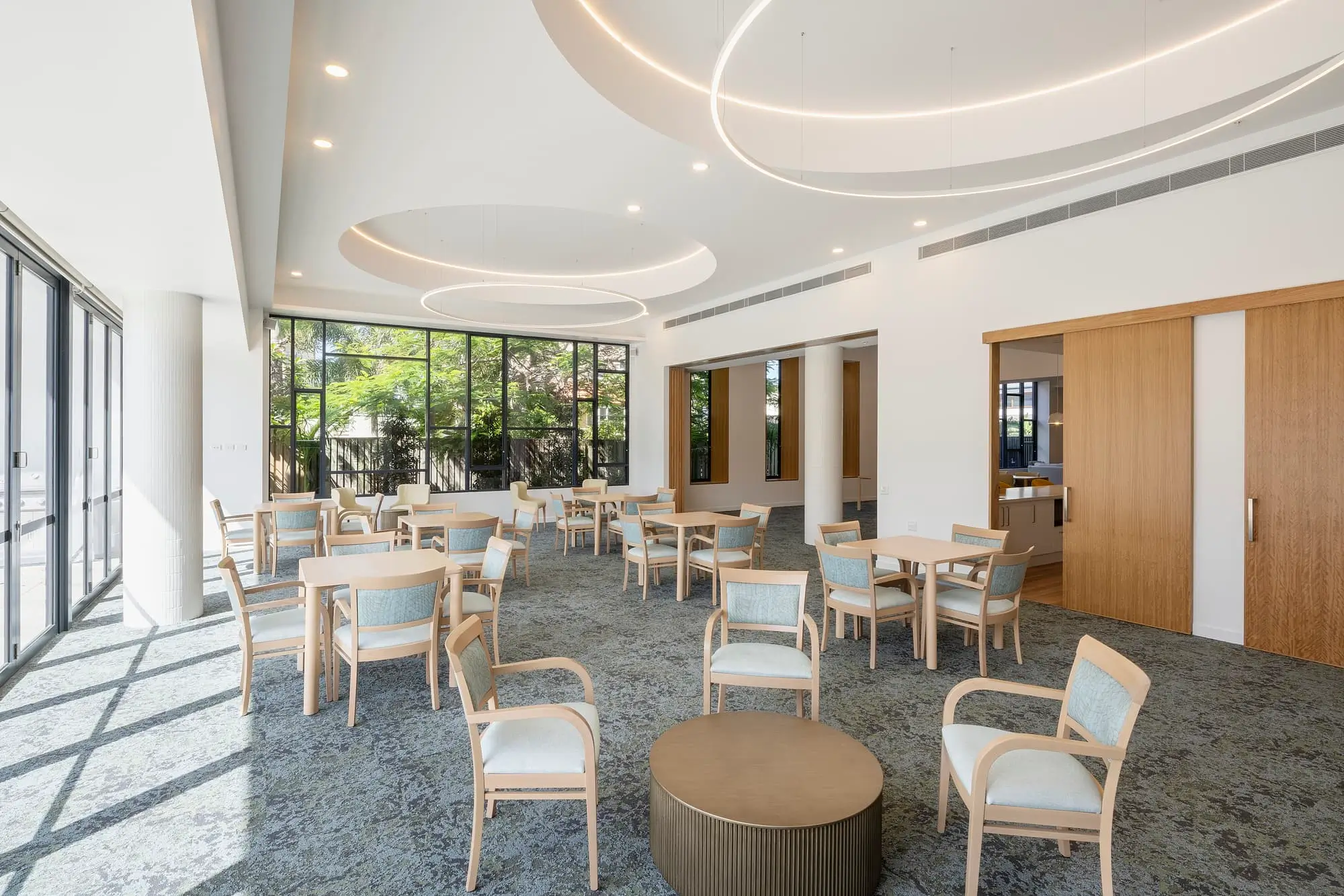 A bright, modern lounge area in this Gold Coast retirement community features round tables and chairs, large floor-to-ceiling windows, circular ceiling lights, and wooden sliding doors. Sunlight streams in, highlighting the clean, open space.