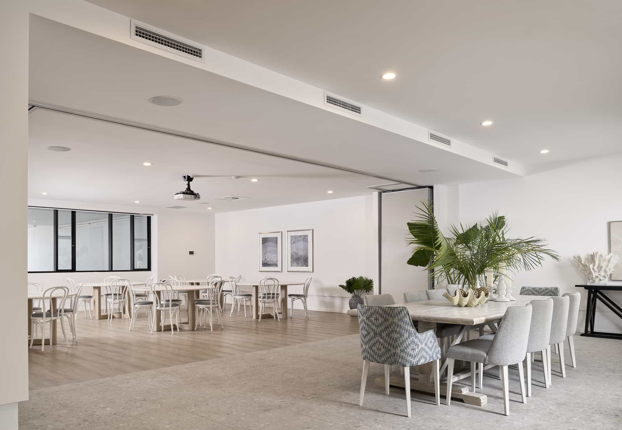 A spacious, modern dining area in the Sunshine Coast Retirement Community features light-colored walls, wooden and tiled floors, white tables and chairs, grey upholstered seating, green plants, framed art, and large windows that fill the space with natural light.