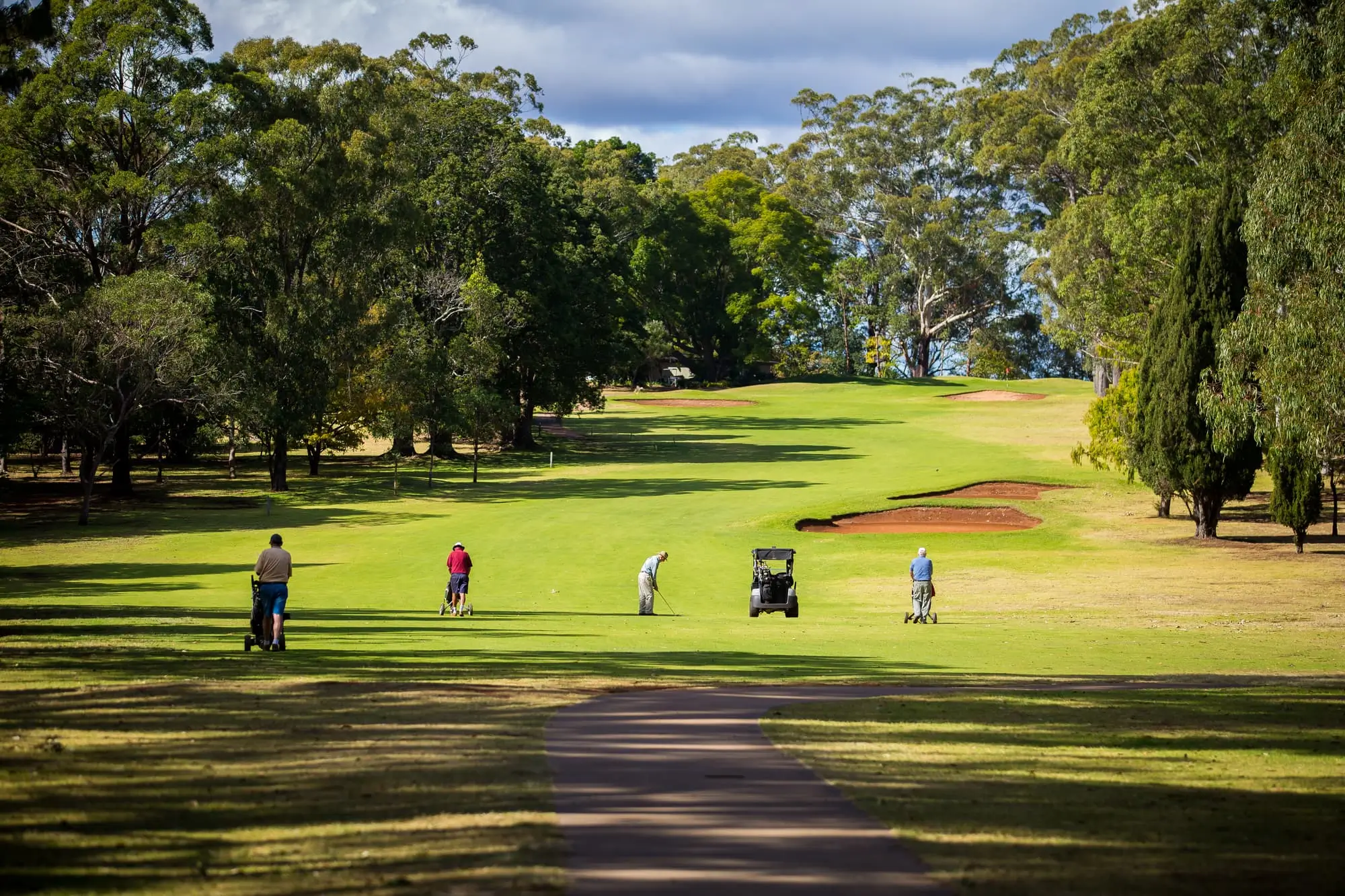 Four people are playing golf on a green course surrounded by trees at the Toowoomba Retirement Community, with a golf cart nearby and sand bunkers visible in the distance under a partly cloudy sky.