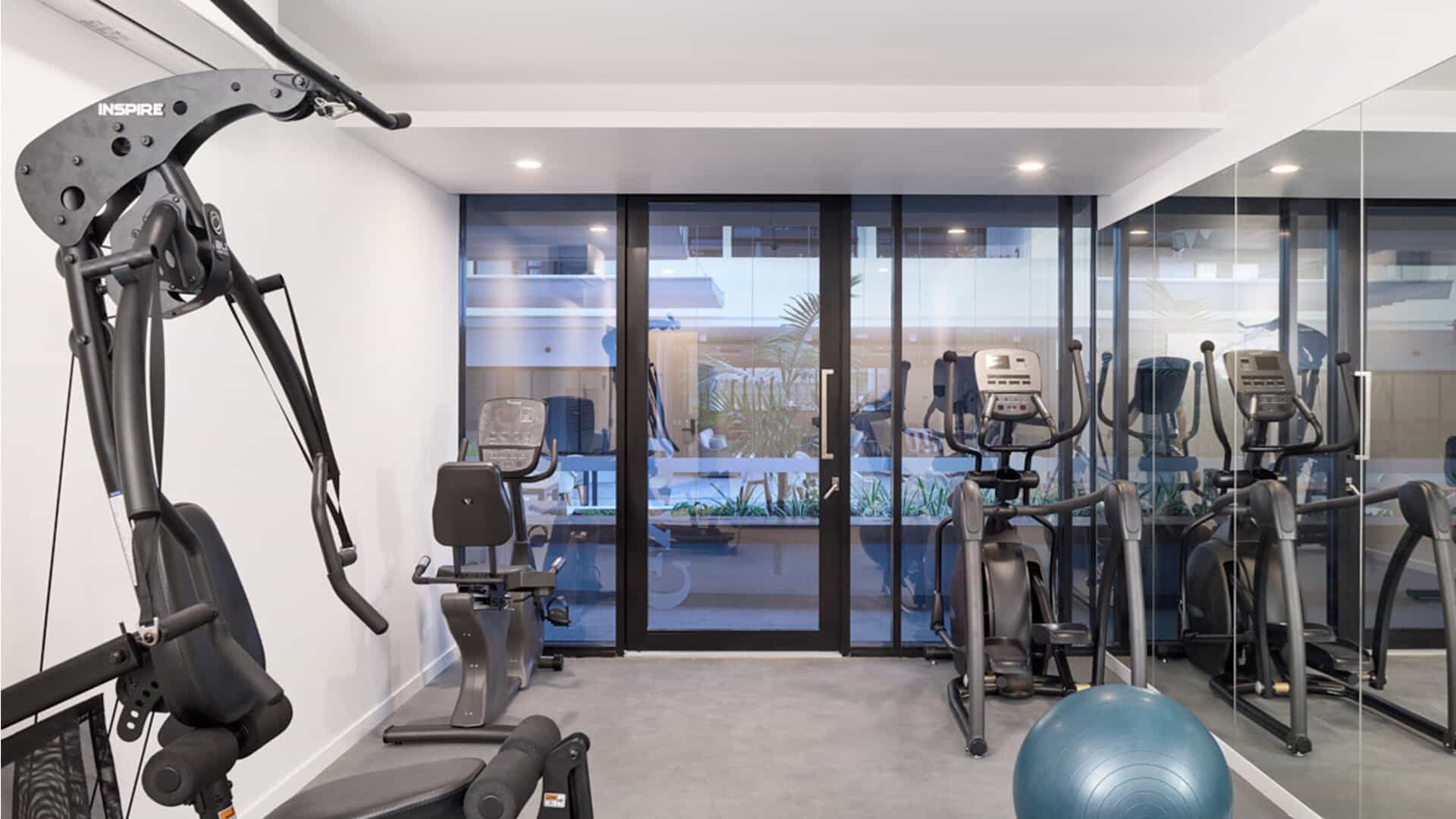 A modern gym room at Sunshine Coast Retirement Community features exercise machines like a multi-gym, recumbent bike, elliptical trainer, and a blue exercise ball. Large windows and mirrored walls create a bright, spacious atmosphere.