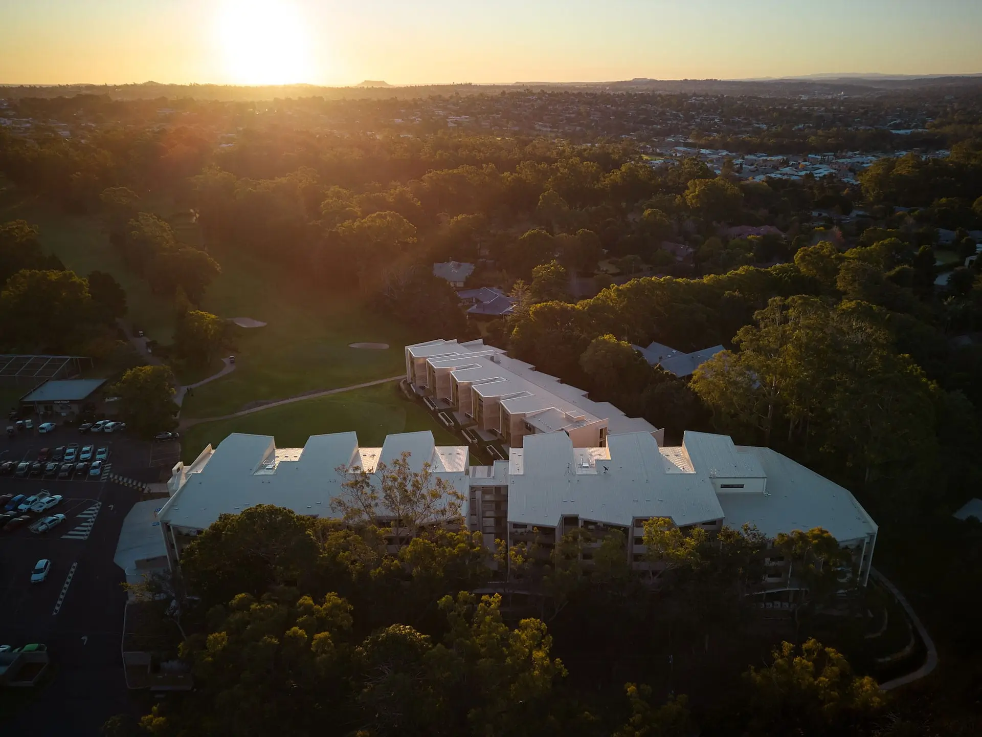 Aerial view of Toowoomba Retirement Community surrounded by trees at sunset, with sunlight casting a warm glow over the landscape and a residential area visible in the background.