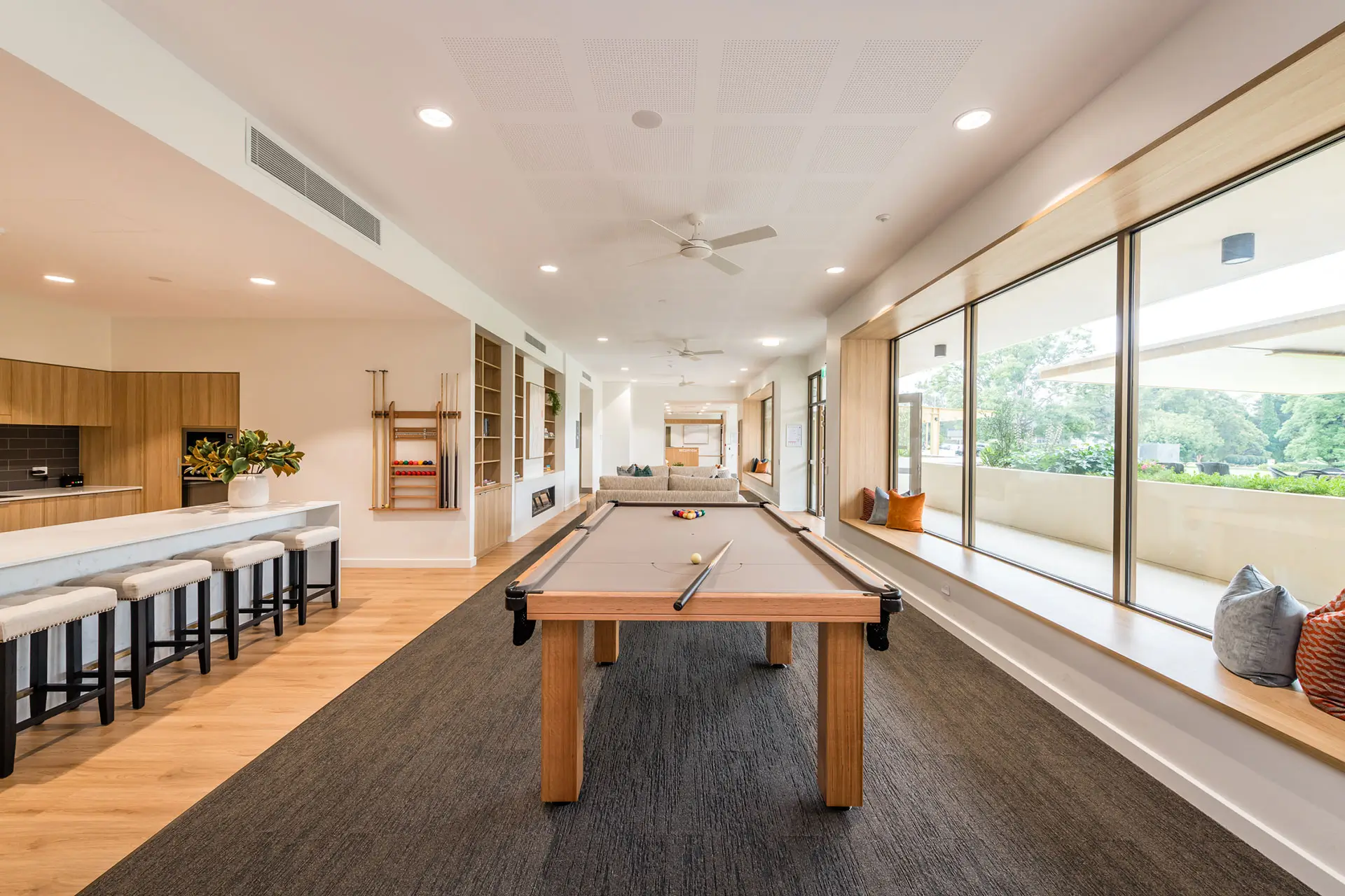 Modern game room at the Toowoomba Retirement Community features a central pool table, bar stools at the kitchen counter, large right-side windows, and built-in shelving with decor. Natural light fills this spacious, stylish area.