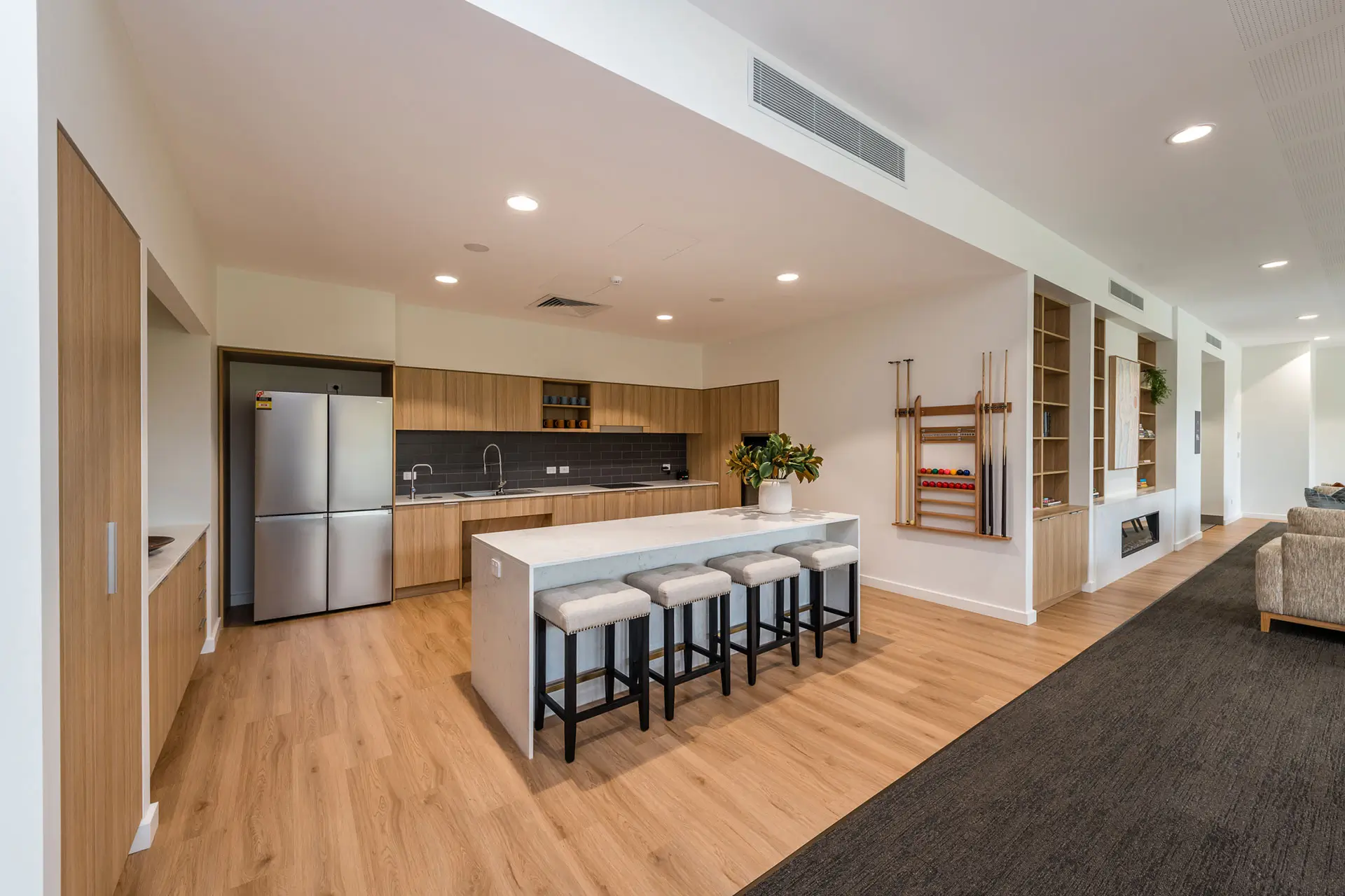 Modern kitchen with wood cabinets, stainless steel refrigerator, island with four padded stools, and open shelving. Perfect for a Toowoomba Retirement Community, it also features a wall-mounted pool cue rack and wine storage. Light wood flooring throughout.