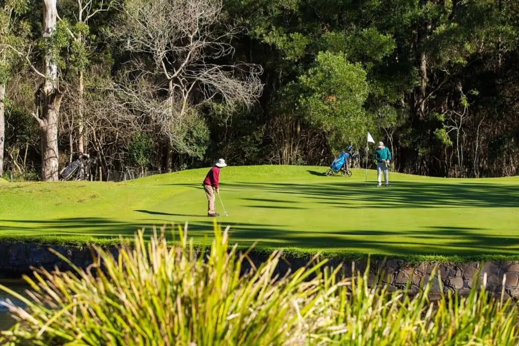 Two people enjoy a game on a golf course at the Toowoomba Retirement Community; one is putting on the green as the other holds the flag near the hole, surrounded by trees, bushes, and sunlight casting gentle shadows across the grass.