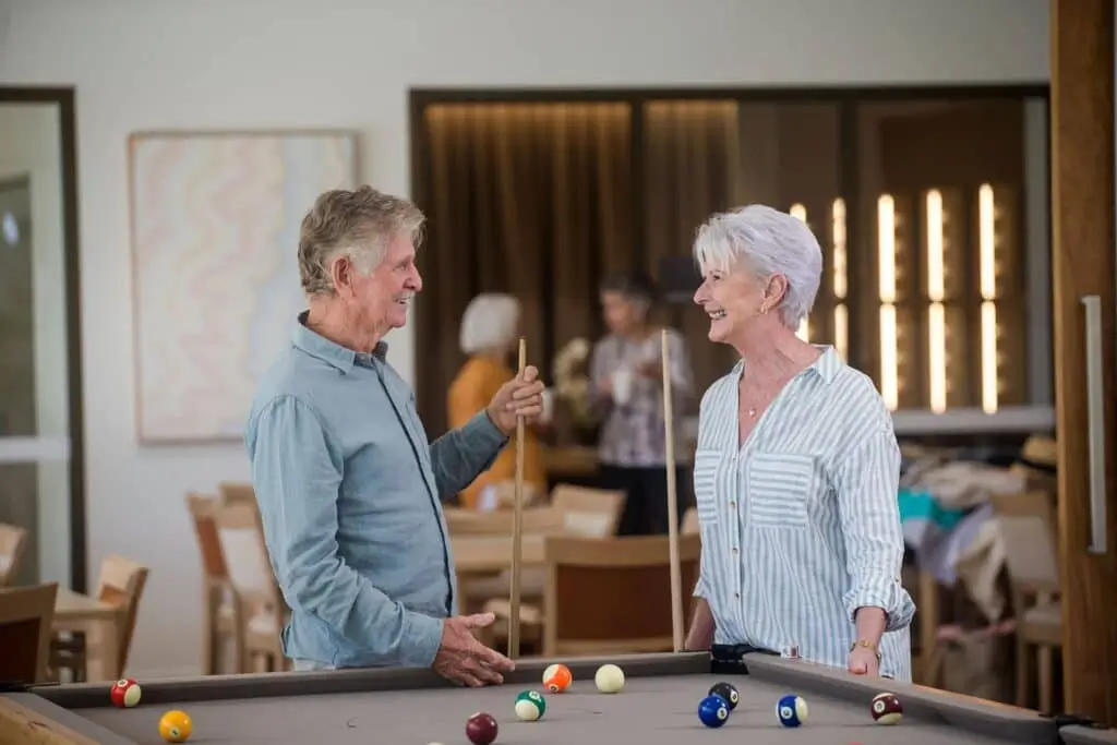 Two smiling older adults stand by a pool table at a Toowoomba Retirement Community, holding cues and chatting. In the background, others gather around tables in a warmly lit room with modern decor.