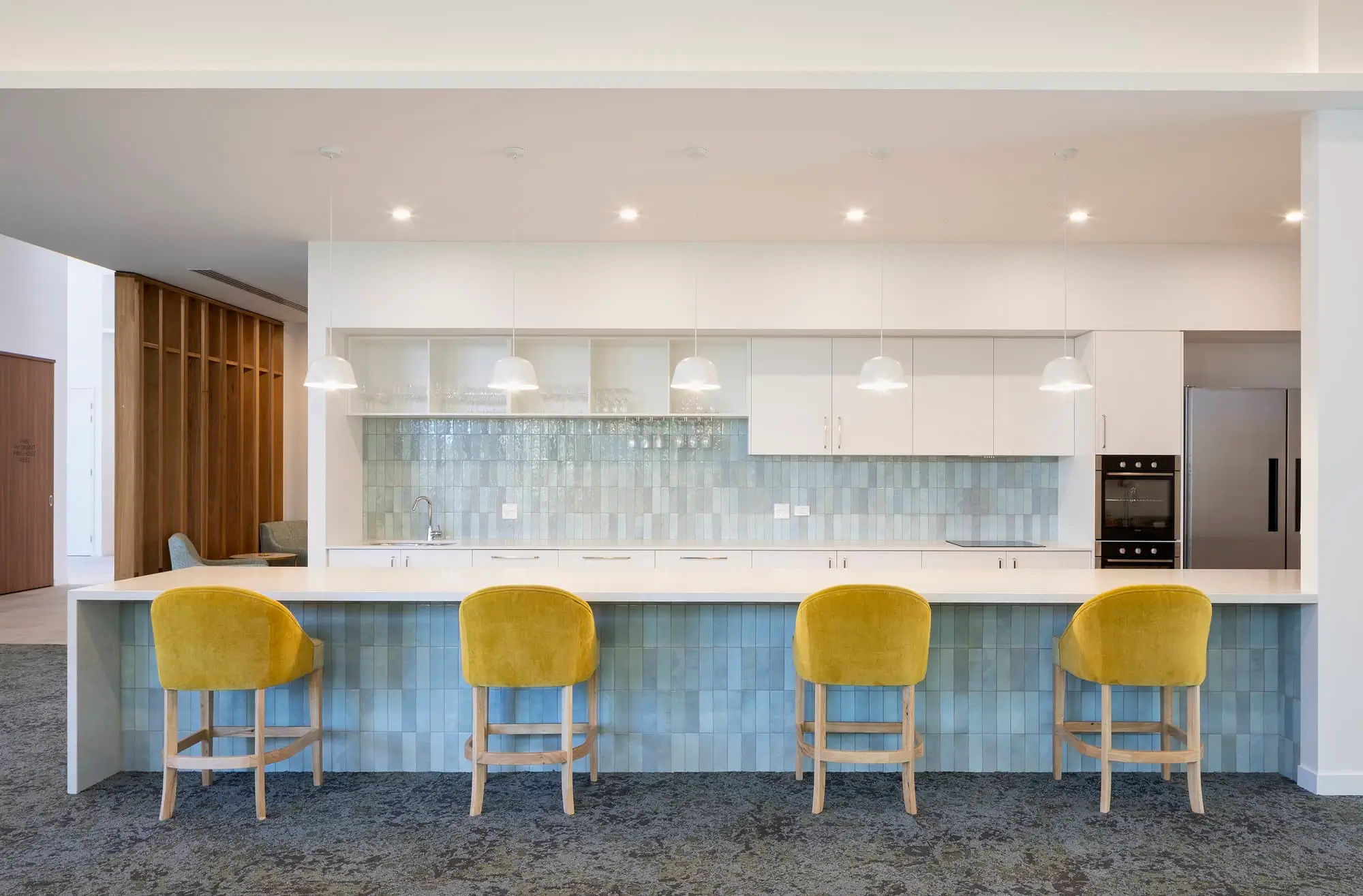 Modern kitchen in a Gold Coast retirement community with a long island, four yellow upholstered bar stools, white cabinets, light blue-green tile backsplash, hanging pendant lights, and built-in appliances.
