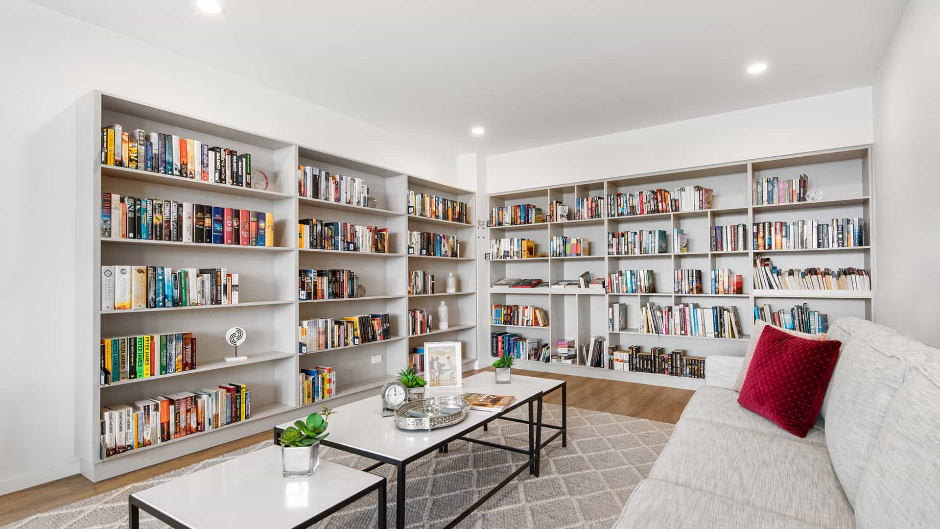 A modern, bright library room in a Sunshine Coast Retirement Community, featuring large bookshelves filled with books, a white sofa with red pillows, two coffee tables with plants and books, and a patterned rug on a wooden floor.