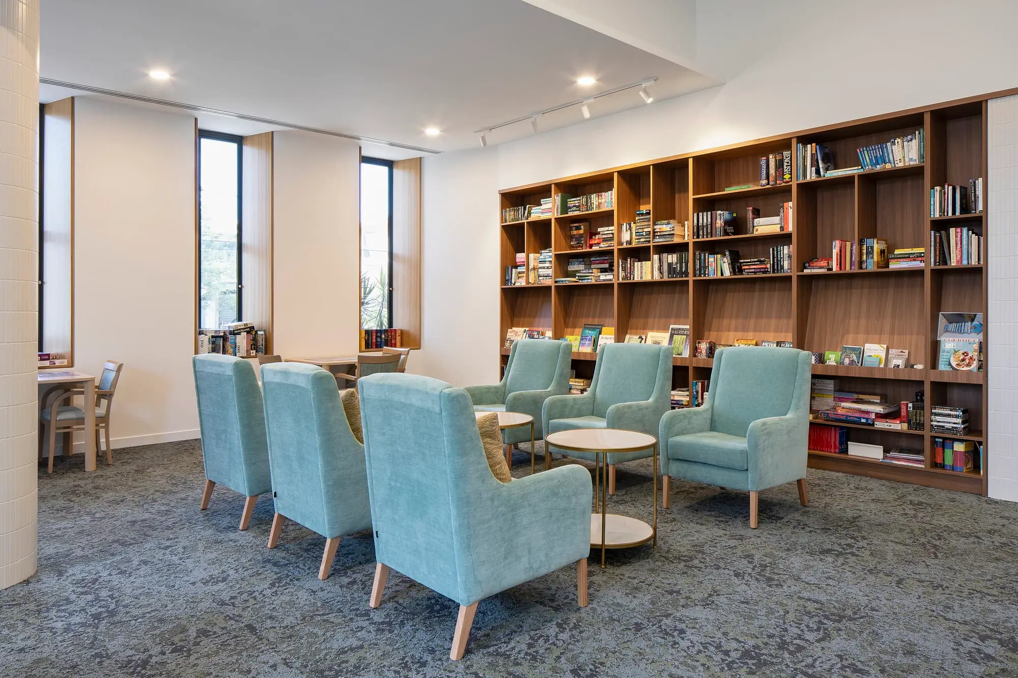 A modern library lounge in a Gold Coast retirement community features light blue armchairs arranged around small round tables, a wall of bookshelves filled with books, and large windows letting in natural light.