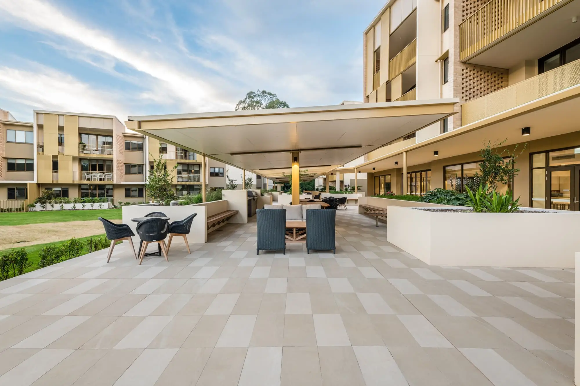 Modern outdoor patio area with lounge chairs and tables under a large canopy, surrounded by landscaped greenery at the Toowoomba Retirement Community, with multi-story apartment buildings in the background on a sunny day.