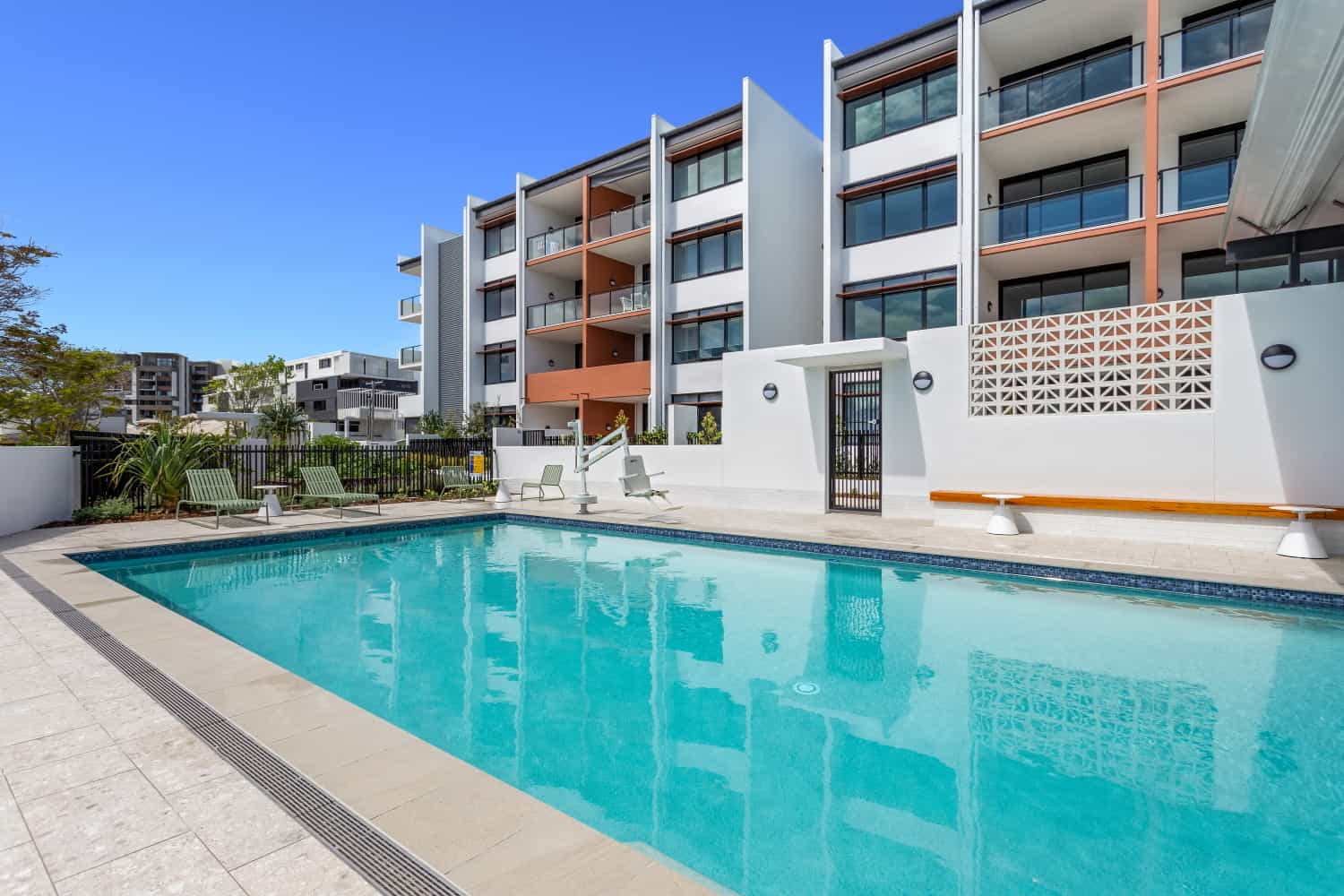 A modern outdoor swimming pool with clear blue water, surrounded by a tiled deck and lounge chairs, is the heart of this Sunshine Coast Retirement Community, with apartments and balconies overlooking the scene under a clear blue sky.