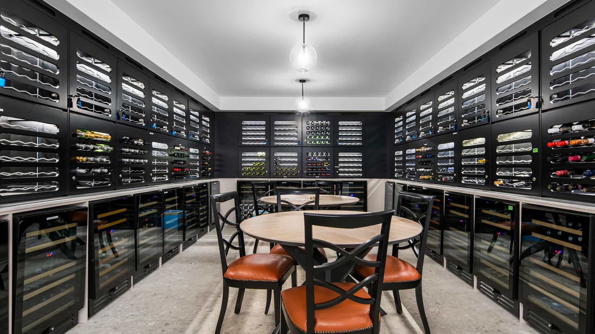 Modern wine cellar with black shelves displaying numerous wine bottles along three walls. A round wooden table with four black chairs sits under pendant lights—an elegant feature often found in exclusive Sunshine Coast Retirement Community residences.