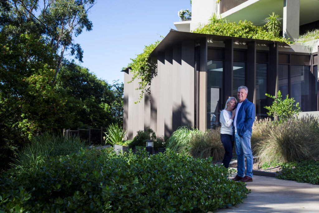 A couple stands together, smiling, in a lush garden outside a modern house with large windows and green plants growing on the building and around the yard. The sky is clear and sunny.