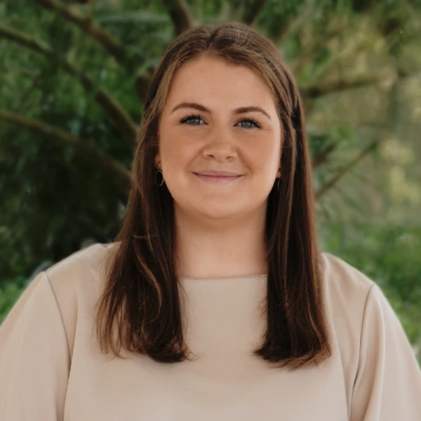 A young woman with long brown hair and blue eyes is smiling softly. She is wearing a beige top and standing outdoors in front of green, leafy trees.