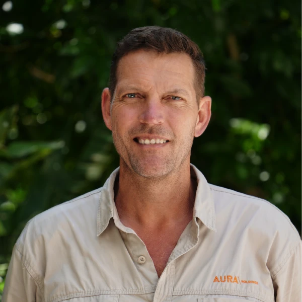 A man with short brown hair wearing a light beige collared shirt with "AURA" embroidered in orange stands outside in front of green foliage, smiling at the camera.