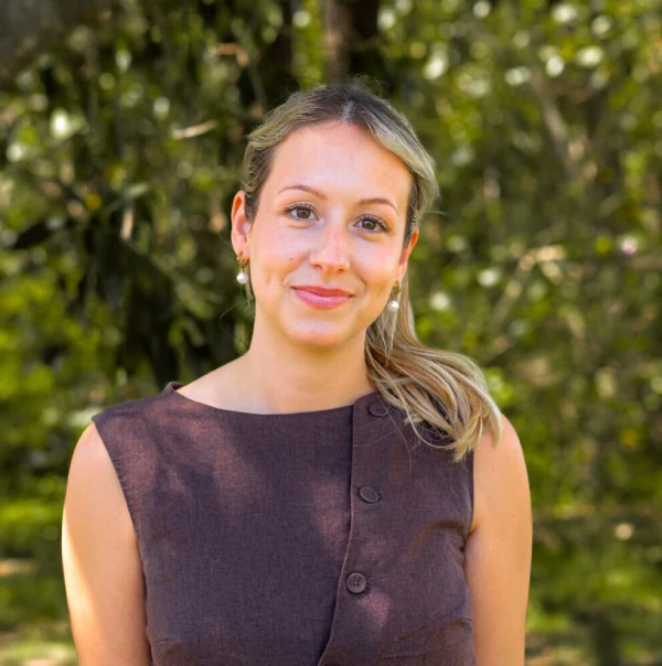 A woman with blonde hair tied back, wearing a sleeveless brown top and pearl earrings, stands outdoors in front of green, leafy trees, smiling softly at the camera.