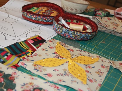 A crafting table with floral fabric, yellow petal-shaped cutouts, colorful fabric strips, scissors, and a sewing box filled with thread spools and sewing supplies. Paper patterns are spread out underneath.