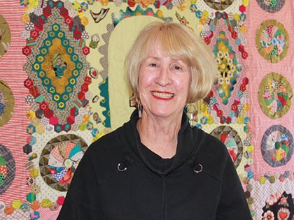 A person with short blonde hair smiles in front of a colorful quilt with various geometric patterns and circular designs. The quilt has a variety of colors, including pink, green, and yellow. They are wearing a black top.