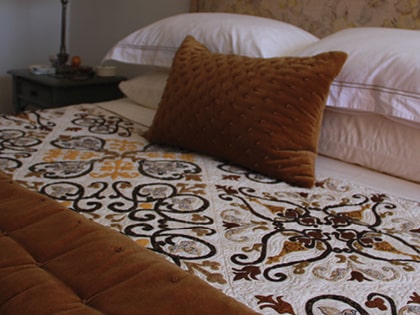 Close-up of a bed with a brown quilted pillow and a patterned bedspread featuring ornate designs in brown, black, and yellow. A side table with a lamp and decorative items is visible in the background.