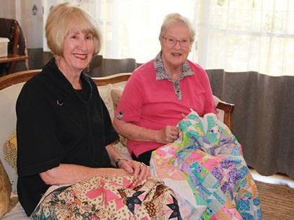 Two elderly women sitting on a couch, each holding a colorful quilt. One wears a black top, the other a pink shirt. They're smiling and appear engaged in conversation. Bright natural light filters through a window behind them.