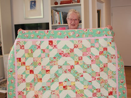 An elderly woman smiles while holding up a colorful patchwork quilt with a pattern of flowers and geometric designs in pink, green, and white. She stands in a cozy room with bookshelves in the background.
