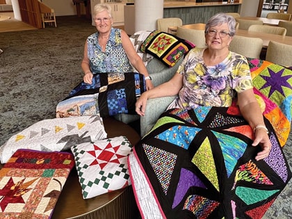 Two women are sitting in a room displaying colorful quilts. They each hold a quilt on their lap, showcasing various intricate patterns and designs. More quilts are spread out on a table beside them, adding to the vibrant display.