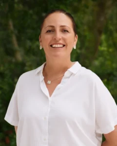 A woman with brown hair tied back, wearing a white button-up shirt, earrings, and a necklace, stands smiling outdoors with a blurred green background.