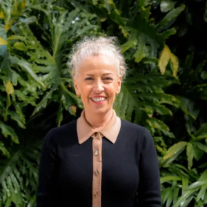 A smiling woman with light hair tied back, wearing a black sweater with a tan collar and gold buttons, stands in front of lush green foliage.