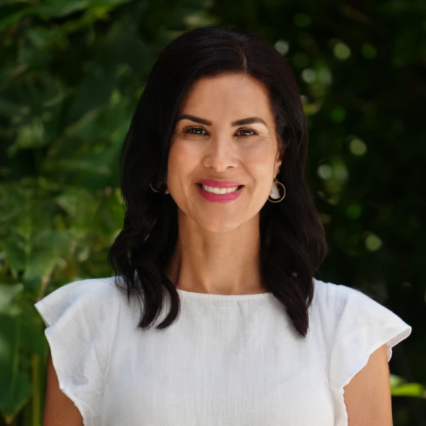 A woman with dark hair and a light complexion smiles at the camera, wearing a white blouse and hoop earrings, standing in front of green foliage.