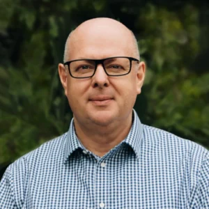 A bald man wearing black glasses and a blue-and-white checkered shirt stands outdoors in front of green, leafy foliage, looking at the camera with a neutral expression.