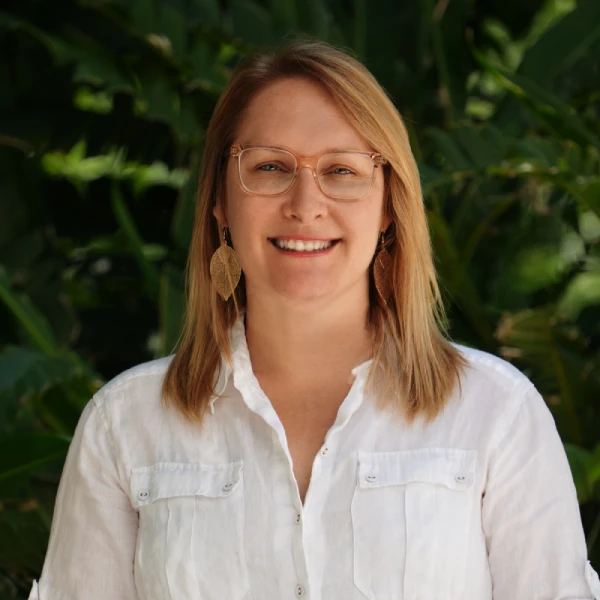 A woman with shoulder-length blonde hair, wearing glasses, leaf-shaped earrings, and a white button-up shirt, smiles while standing in front of green leafy plants.