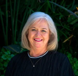 A smiling older woman with short gray hair wearing a black top and necklace stands in front of lush green plants.