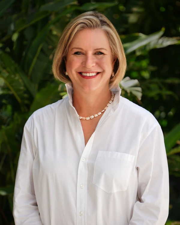 A woman with short blonde hair smiles, wearing a white collared shirt and a beaded necklace, standing outdoors in front of green leafy plants.