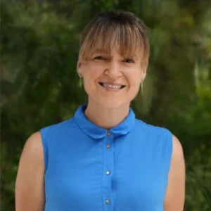A woman with light brown hair and bangs smiles at the camera, wearing a sleeveless blue button-up shirt. The background is blurred greenery.