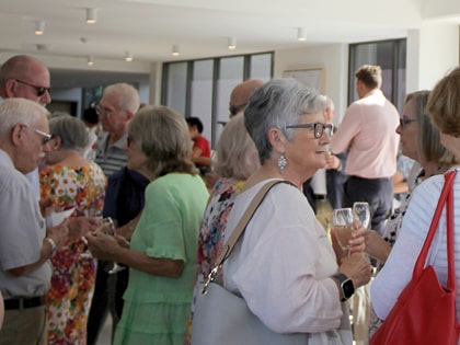 A group of older adults are socializing in a well-lit room. They are engaged in conversation, holding drinks, and wearing casual attire. Some individuals have glasses and there is a mix of colorful and neutral clothing.