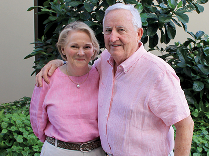 An elderly couple stands together, both wearing pink shirts. The man has his arm around the woman. They are smiling and surrounded by green foliage.