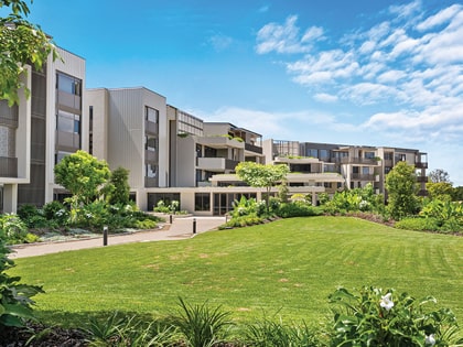 A modern apartment building with multiple stories is surrounded by lush green landscaping under a bright blue sky. A well-maintained lawn and various plants enhance the scenic view.