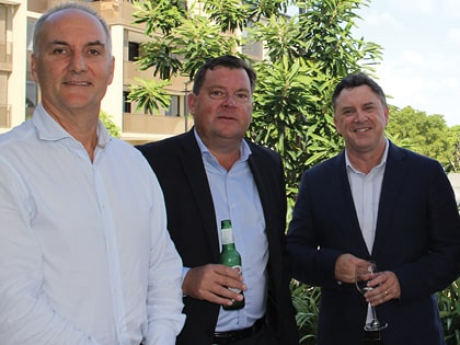 Three men in business attire stand outdoors. One holds a beer bottle, another holds a wine glass. They are surrounded by greenery and modern architecture.