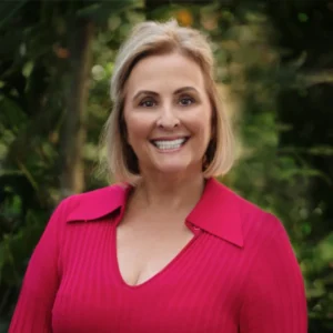 A woman with blonde hair wearing a bright pink blouse smiles outdoors in a lush, green garden setting.