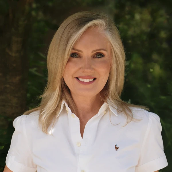 A woman with blonde hair wearing a white collared shirt stands outdoors in front of greenery, smiling at the camera.