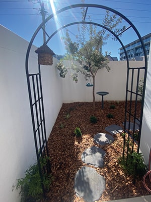 A garden with a wooden archway, circular stepping stones, and small plants is surrounded by white walls. There's a birdbath and a hanging woven decoration. Sunlight shines on the greenery, and a building is visible in the background.