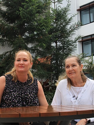 Two women sitting at a wooden table outdoors, surrounded by lush greenery and modern buildings. One woman wears a sleeveless black top with white patterns, and the other is in a white shirt. Both are smiling and appear relaxed.