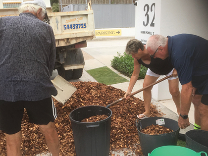 Three people are shoveling bark chips from a pile into black bins in a driveway. They are standing near a white wall with the number 32 on it. A white truck with writing on it is parked nearby.