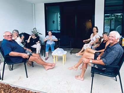 A group of eight people sitting outside on a patio, enjoying drinks and snacks. They are seated in a semi-circle on chairs with a small table in the center. The background shows a building with large windows.