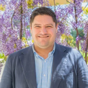A man in a blue blazer and checkered shirt smiles in front of hanging purple wisteria flowers outdoors on a sunny day.