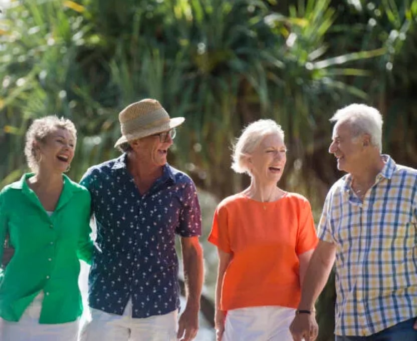 Four older adults, two men and two women, walk outdoors together, smiling and laughing. They are casually dressed and holding hands, with greenery in the background.