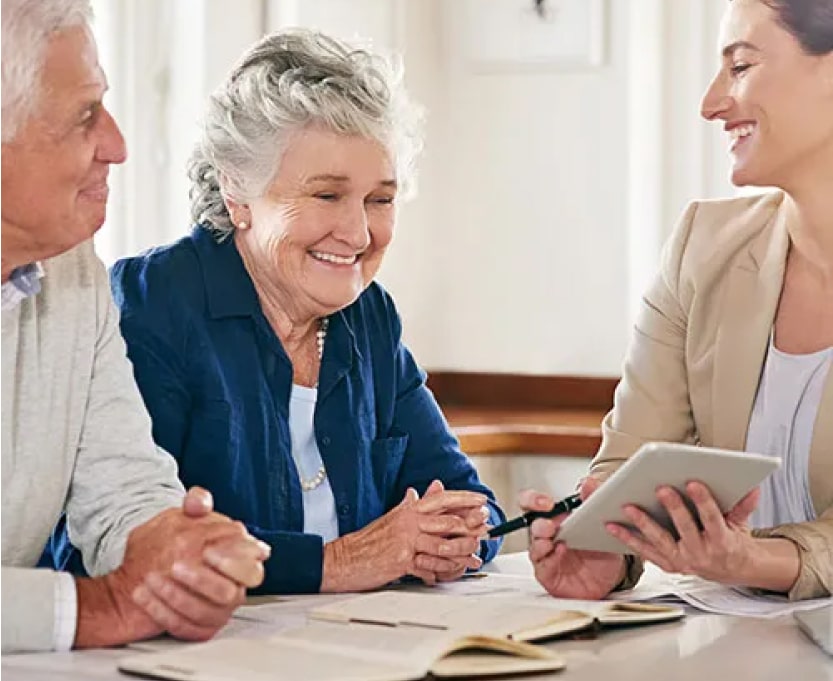 Two smiling older adults sit at a table with a younger woman, who is holding a tablet and pointing at it while discussing documents with them in a bright, indoor setting.