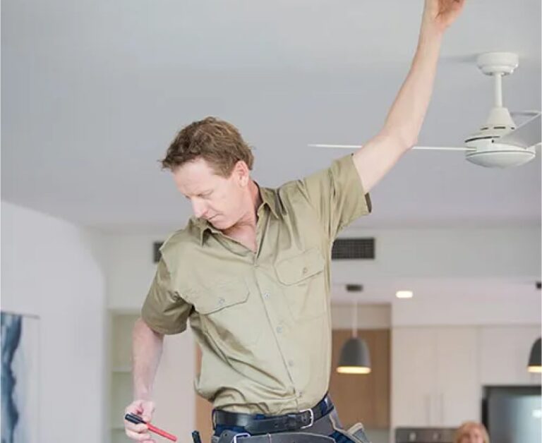 An electrician in a khaki shirt stands on a ladder adjusting a ceiling fan with one hand, holding tools in the other, in a modern home interior.